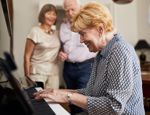 Older lady playing the piano with friends listening behind.