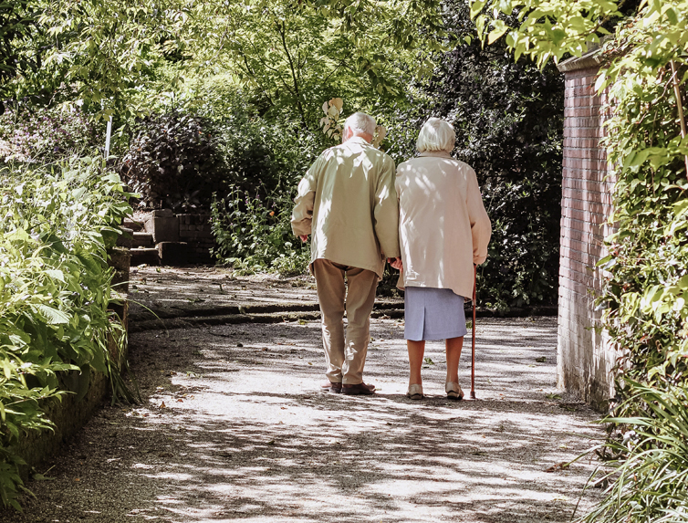 Older couple walking together in dappled light and greenery.